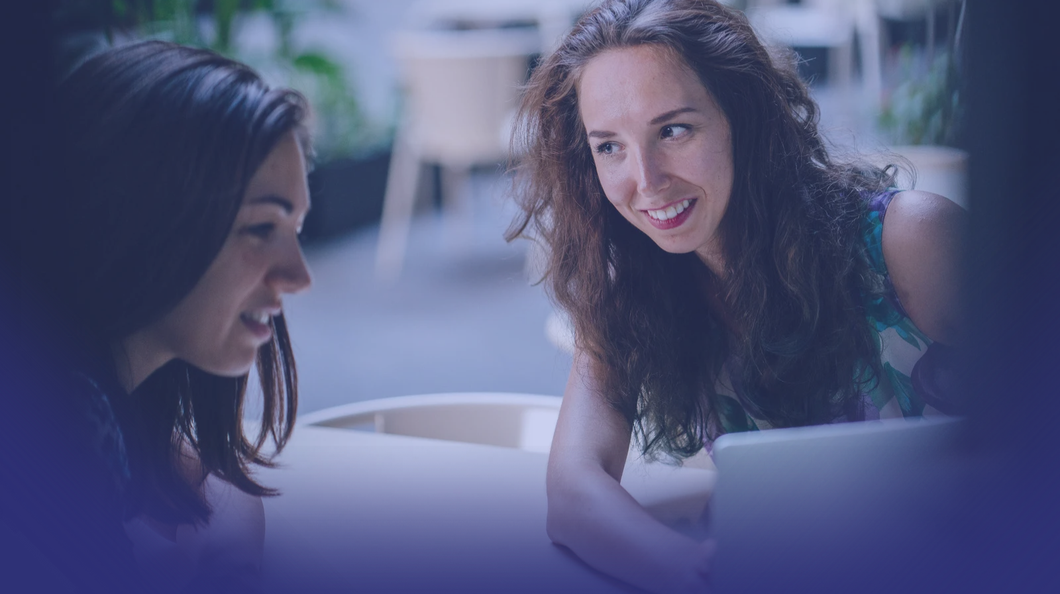 Two women smiling and having a conversation at a table, with a blurred outdoor background.