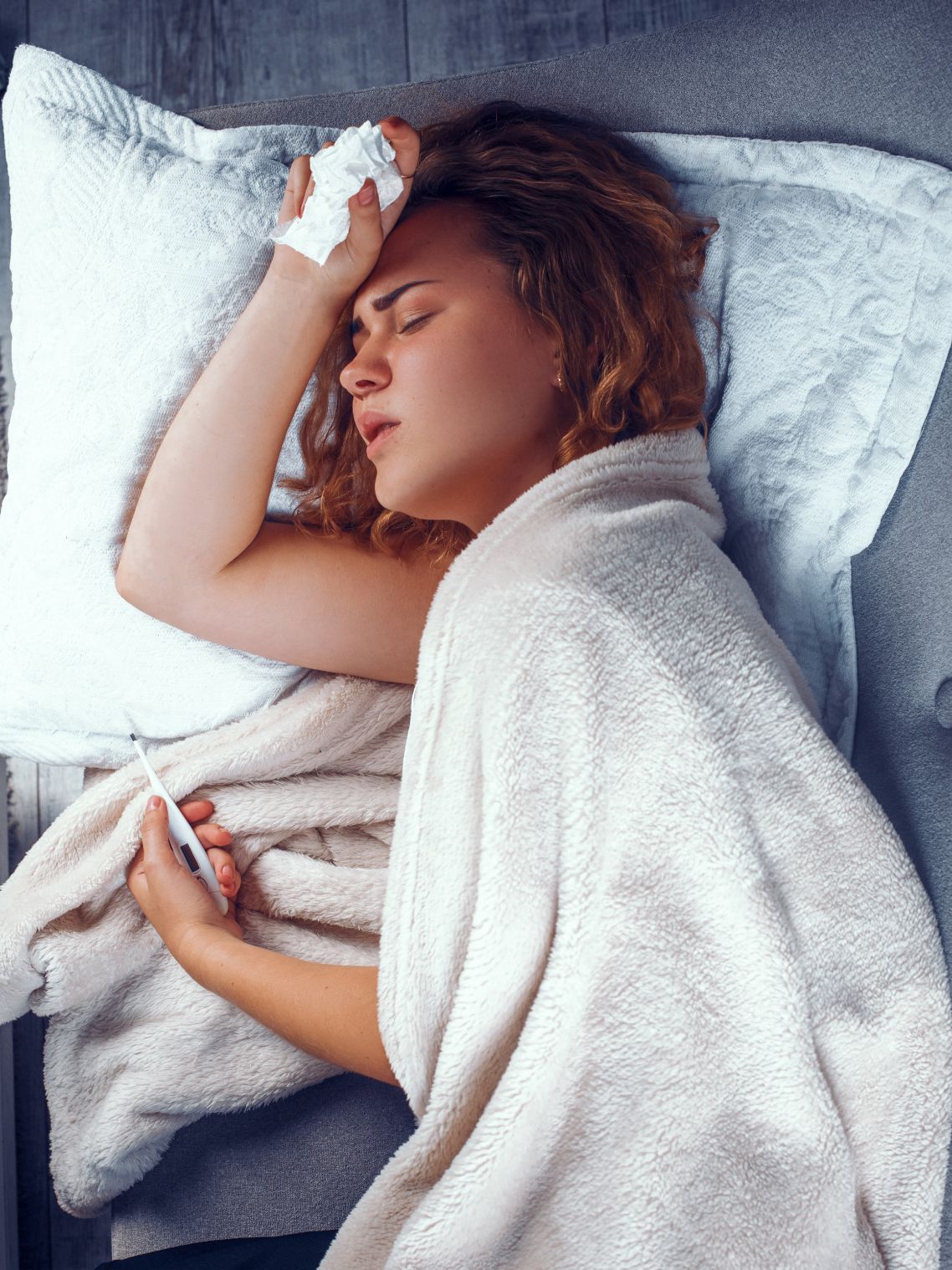 Eine Frau liegt krank im Bett, hält sich den Kopf und in der anderen Hand ein Fieberrthermometer.