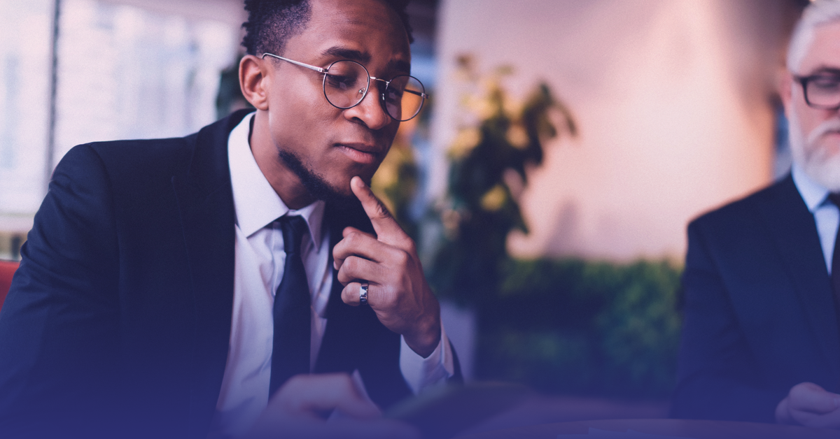 Professional man in glasses thoughtfully reviewing something, a colleague partially visible beside him.