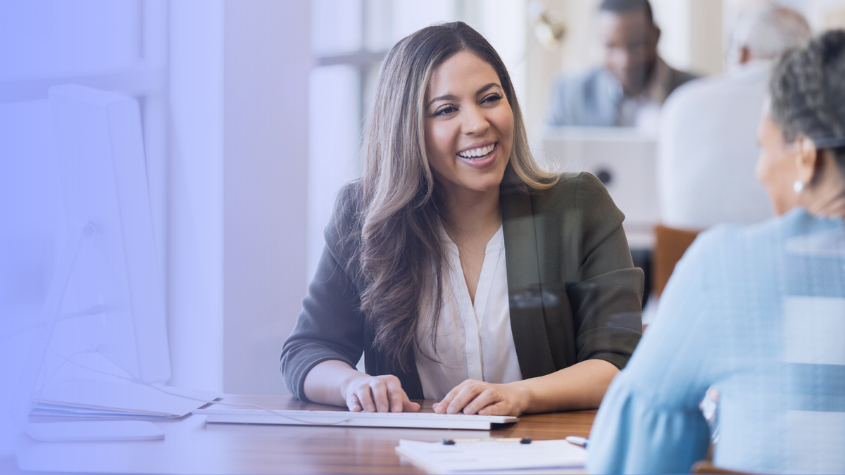 A woman sitting at a desk, smiling and talking to another person in an office setting with people in the background.
