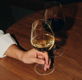 Hand with a glass of white wine at a table, with a glass of red wine in the background.