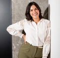 Vrouw met donker haar glimlacht in witte blouse met kant en groene broek, leunend tegen een marmeren muur.