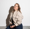 Vrouw zittend op een bank met een mok in haar hand, draagt een beige jas en blauwe broek, tegen een witte achtergrond.