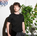 Man met krullend haar houdt een laptop vast in een kamer met planten, een bokaal en een basketbalnet op de achtergrond.