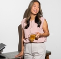 Maria in een roze blouse en grijze broek leunt lachend tegen een tafel, met een glas thee in haar hand.