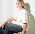 Persoon met kort blond haar lacht, zittend op een stoel. Ze draagt een wit T-shirt en een spijkerbroek. Schaduw op witte muur.