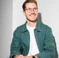 Man met bril glimlacht, draagt een groene jas en witte T-shirt, handen gevouwen op schoot, zittend tegen een witte muur.