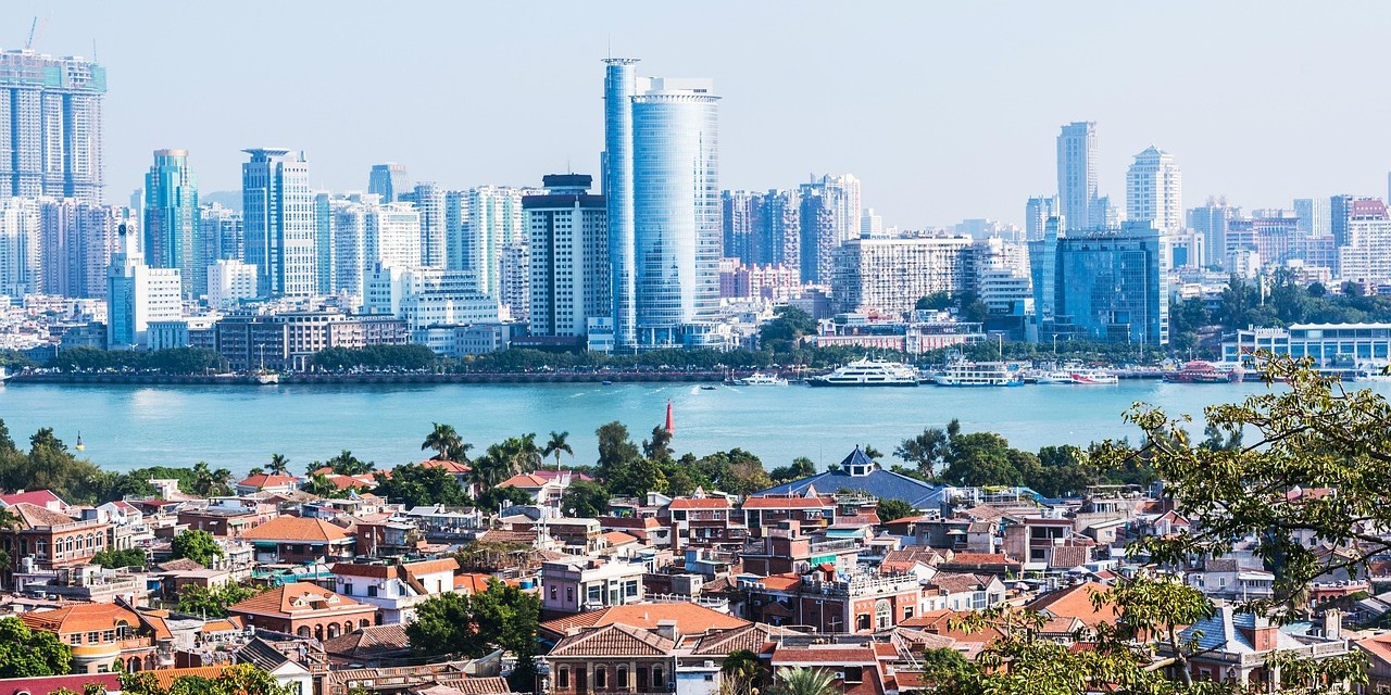 The image shows a cityscape with modern skyscrapers across a river, traditional red-roofed buildings in the foreground, and greenery on a sunny day.