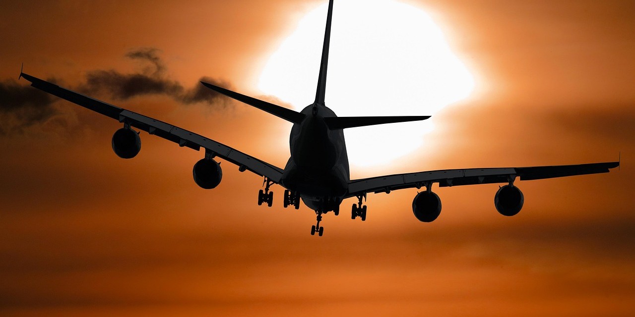 Silhouette of a large airplane flying against a dramatic orange sunset sky, with the sun glowing behind the aircraft.