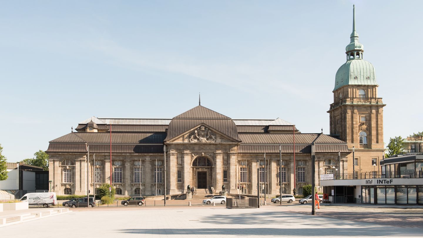 Historisches Gebäude mit gewölbtem Eingang und großem Kuppelturm auf der rechten Seite, vor klarem blauen Himmel, mit einem Platz im Vordergrund.