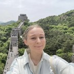 A person takes a selfie with the Great Wall of China in the background. The scene includes lush green hills and a historic watchtower.