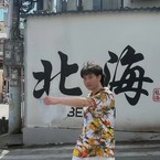 A person wearing a colorful floral outfit stands on a crosswalk, motioning forward, in front of a wall with large Chinese characters and "Be" visible.