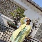 A person in a light green floral dress and yellow coat shields their eyes from the sun, standing by a decorative wall and potted plants.