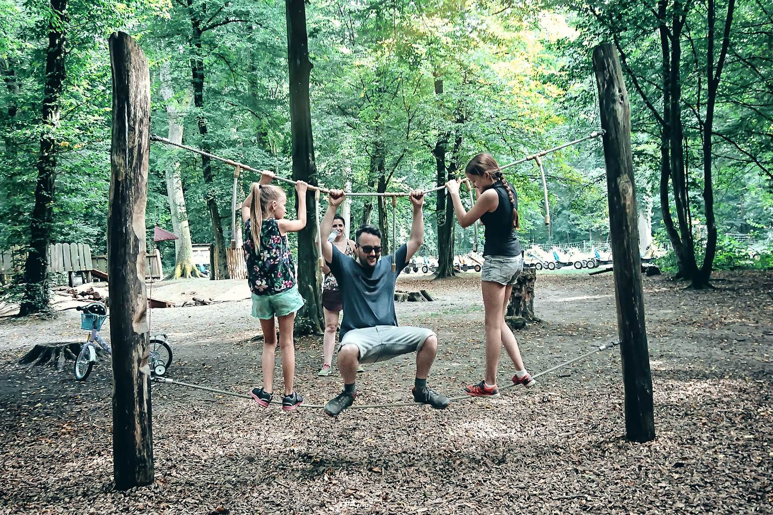 A father balances on a rope with his two daughters in an outdoor playground.