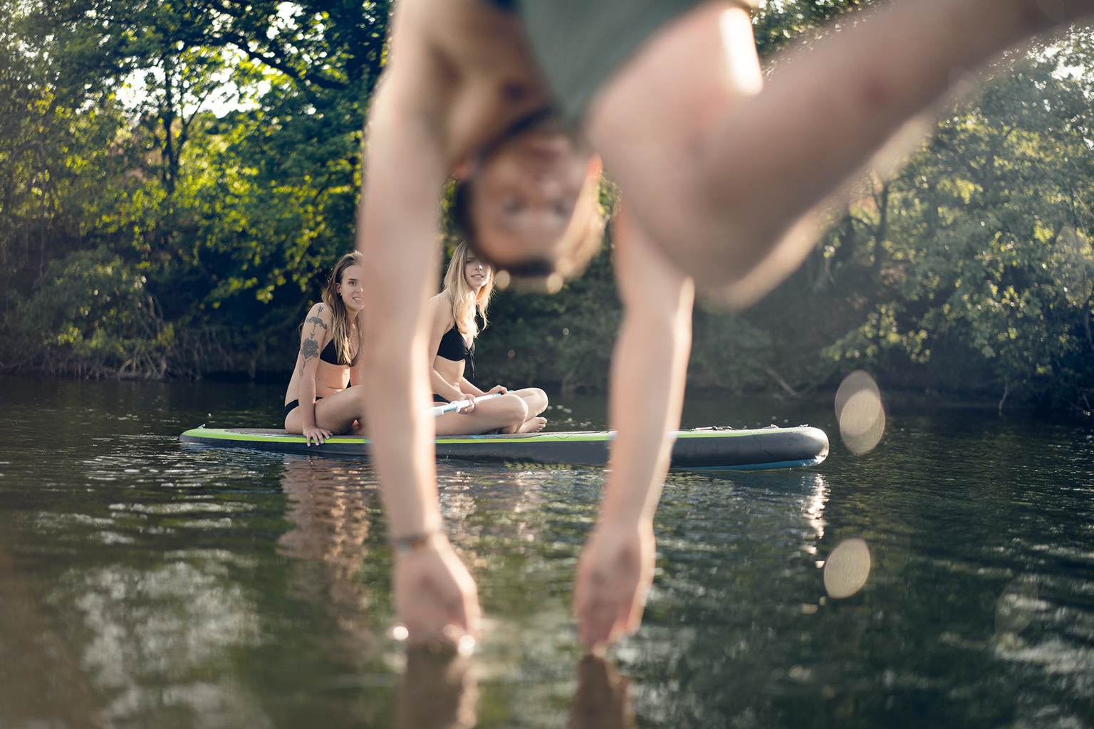 Ein Mann springt mit einem Köpfer in einen Fluss. Im Hintergrund sitzen zwei Frauen auf einem Paddleboard.