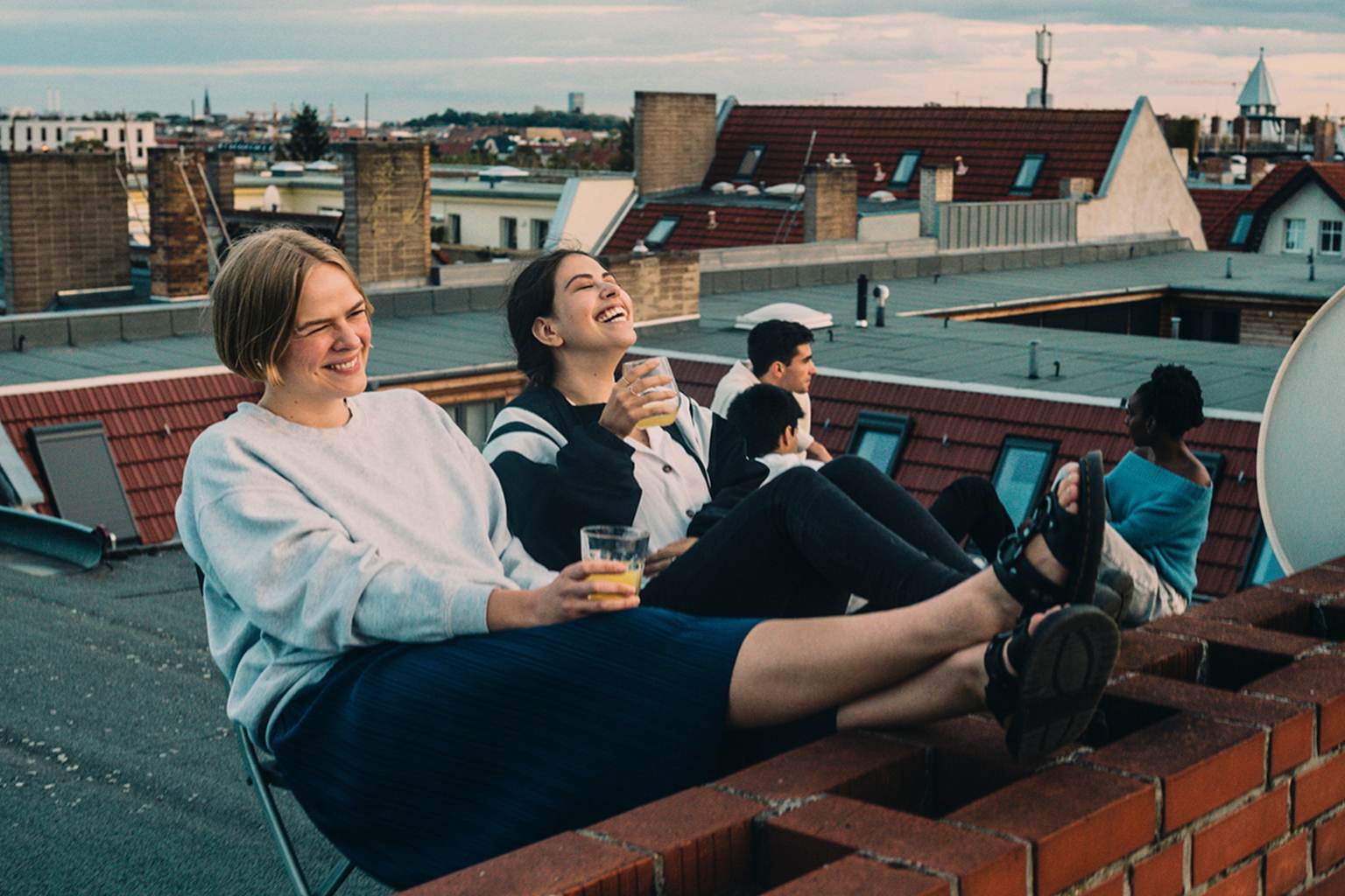 Two women sit laughing on a flat roof. They are both holding a drink in their hands. Three other people are sitting in the background.