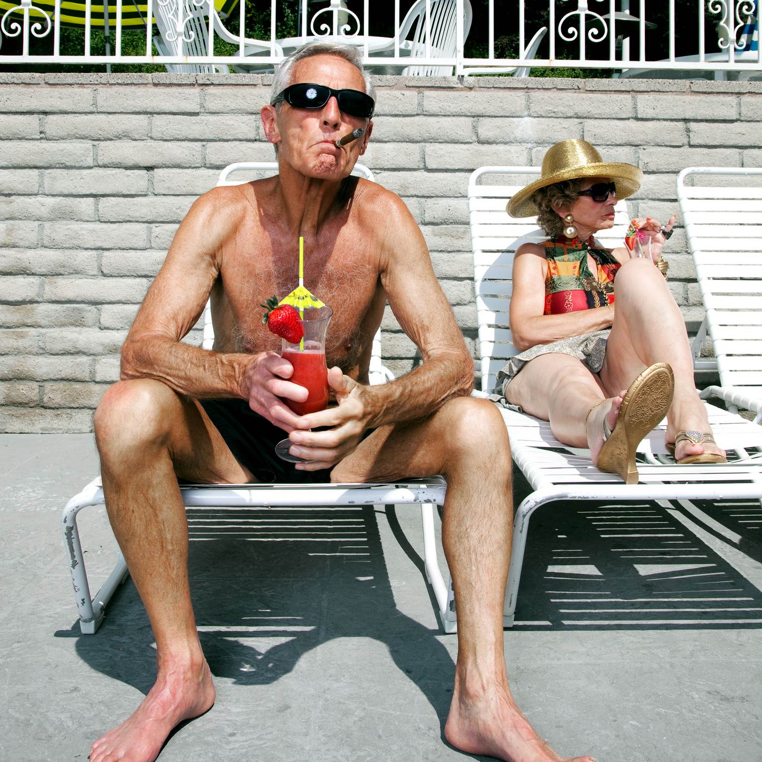 Ein älterer Mann und eine ältere Frau sitzen in Schwimmkleidung auf Liegestühlen. Beide halten einen Cocktail in der Hand. Der Mann raucht eine Zigarre.