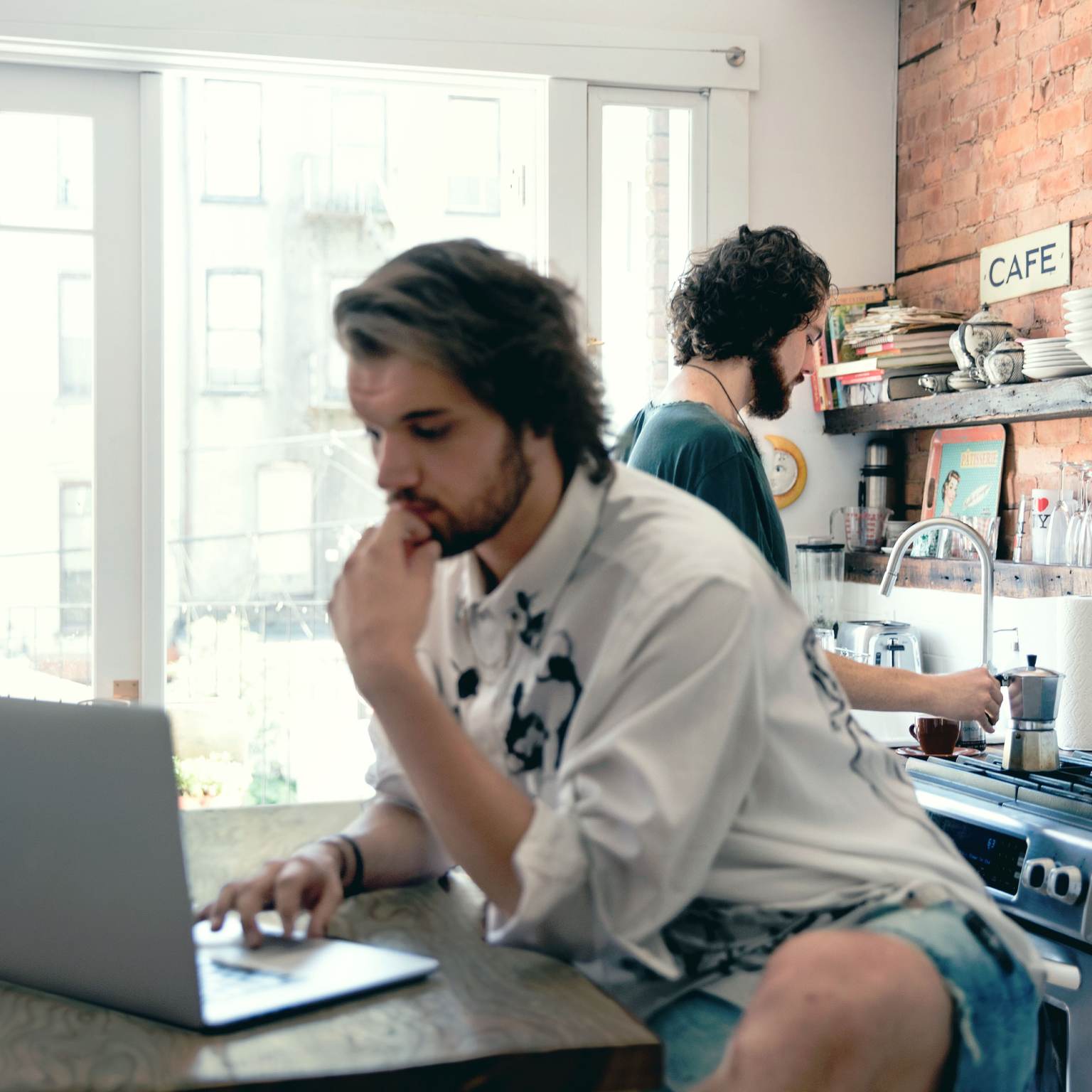 A man is sitting at a table working on a laptop. In the background, another man is making coffee.