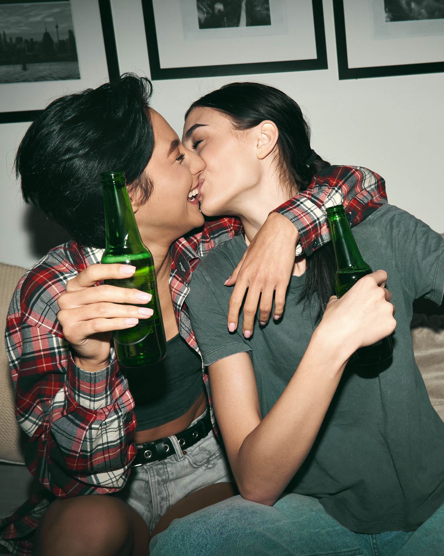 Two women are sitting on a couch and kissing. Both are holding a beer bottle.