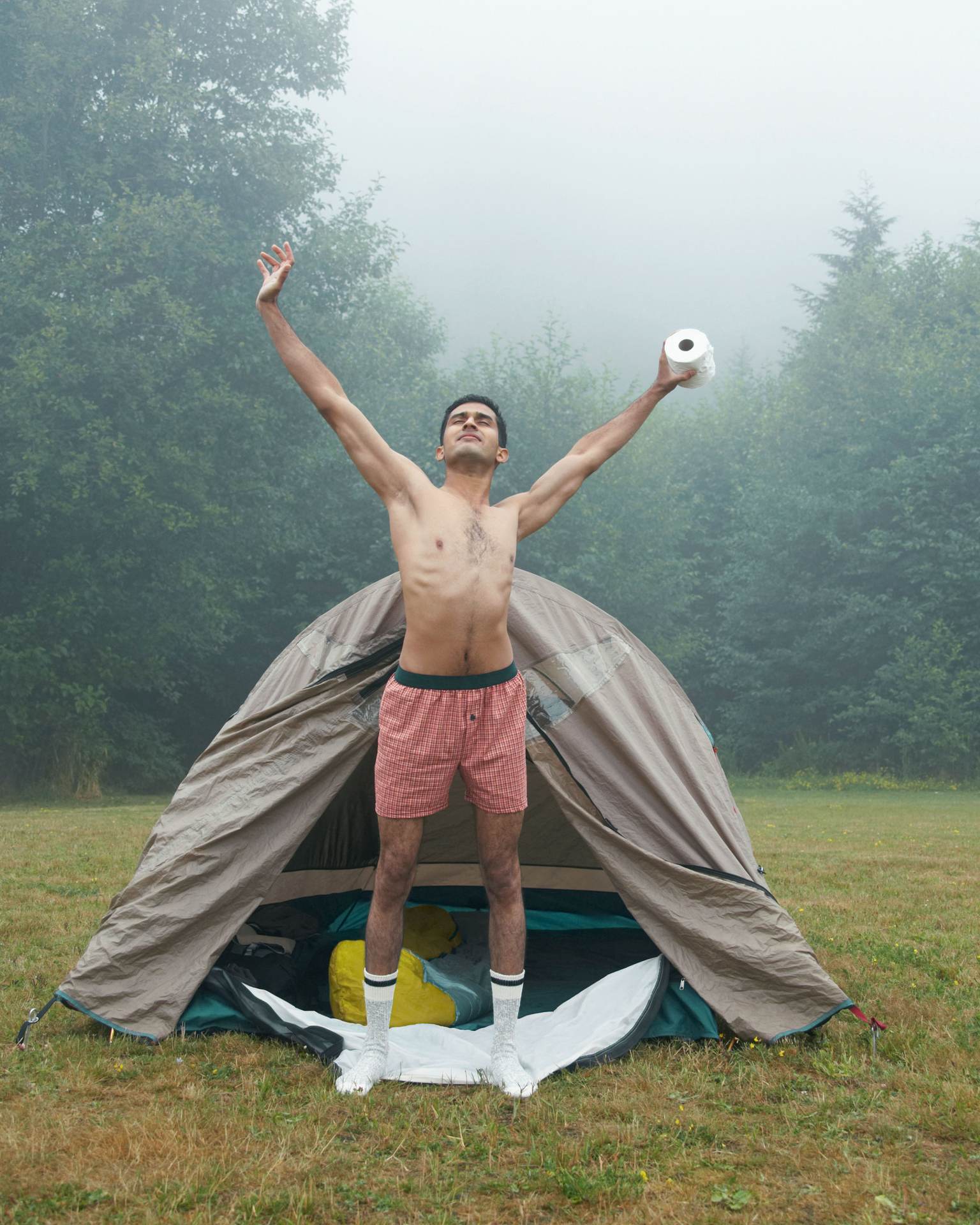 A man in swimming trunks stands in front of a tent and stretches. In his left hand he holds toilet paper.