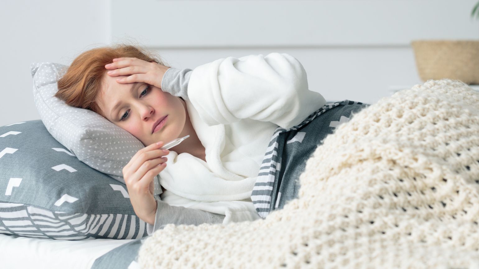 Eine Frau liegt im Bett, hält sich die linke Hand an die Stirn und hält in der rechten Hand ein Fieberthermometer.