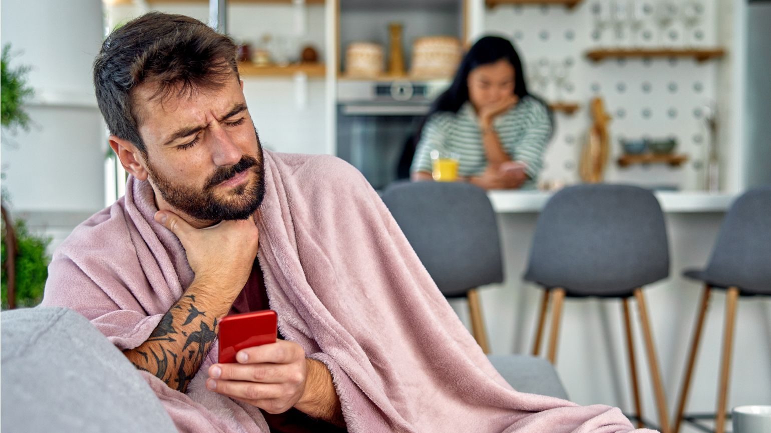 Ein Mann hält in der linken Hand ein Mobiltelefon und fasst sich mit der rechten Hand an den Hals. Im Hintergrund sieht man eine Frau, die an einem Küchentisch sitzt.