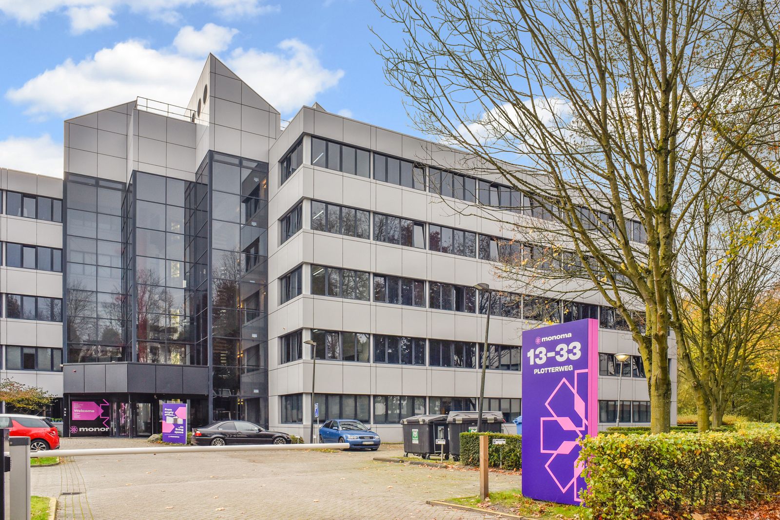 Modern office building with large glass facade, several parked cars, and a purple sign displaying numbers 13-33, surrounded by trees.