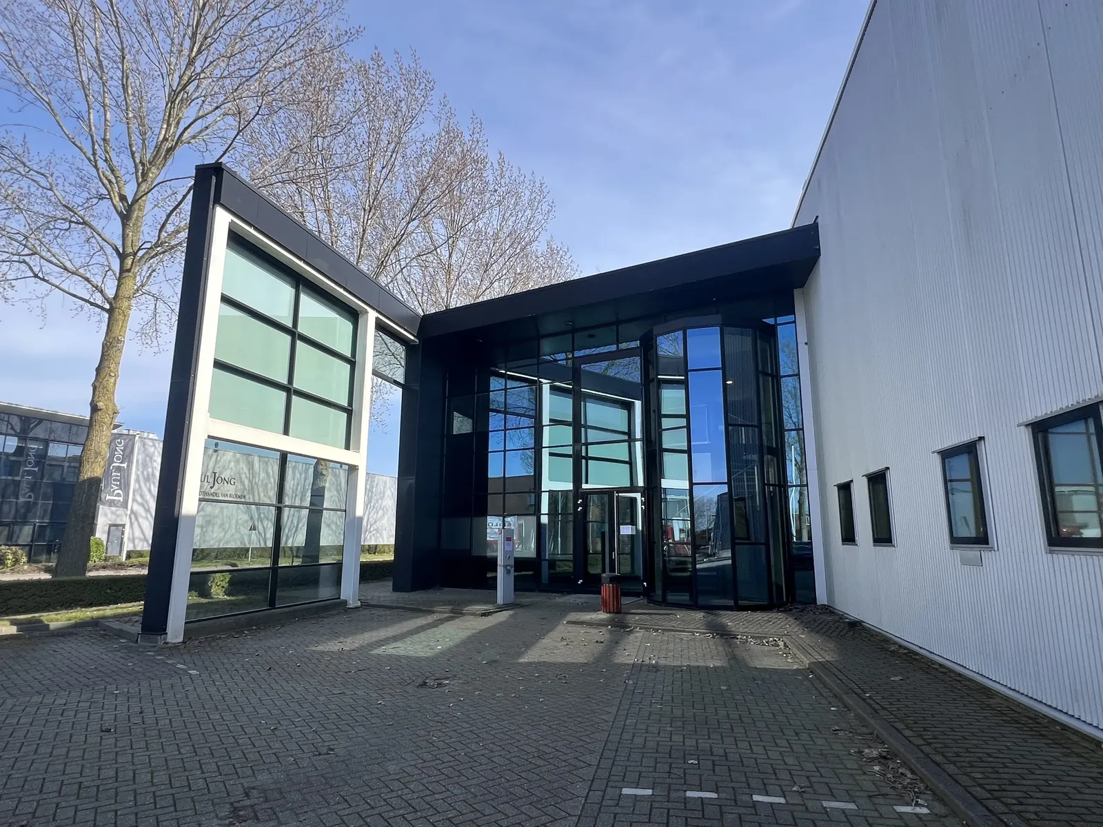 Modern building entrance with large glass panels and black framing, surrounded by pavement and bare trees under a clear blue sky.