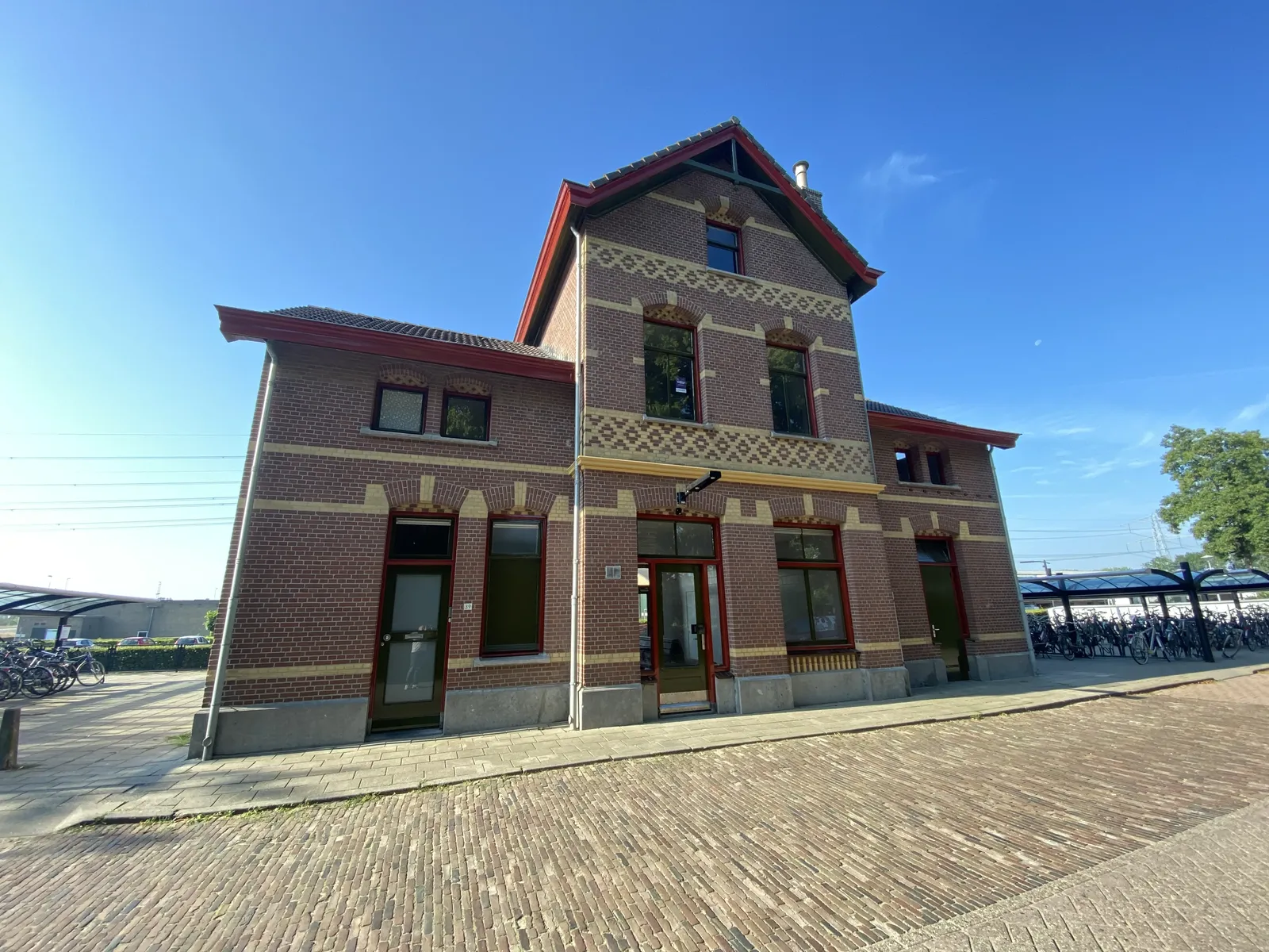 Historic brick railway station building with decorative patterns under a clear blue sky, surrounded by a cobblestone path and bicycle racks.