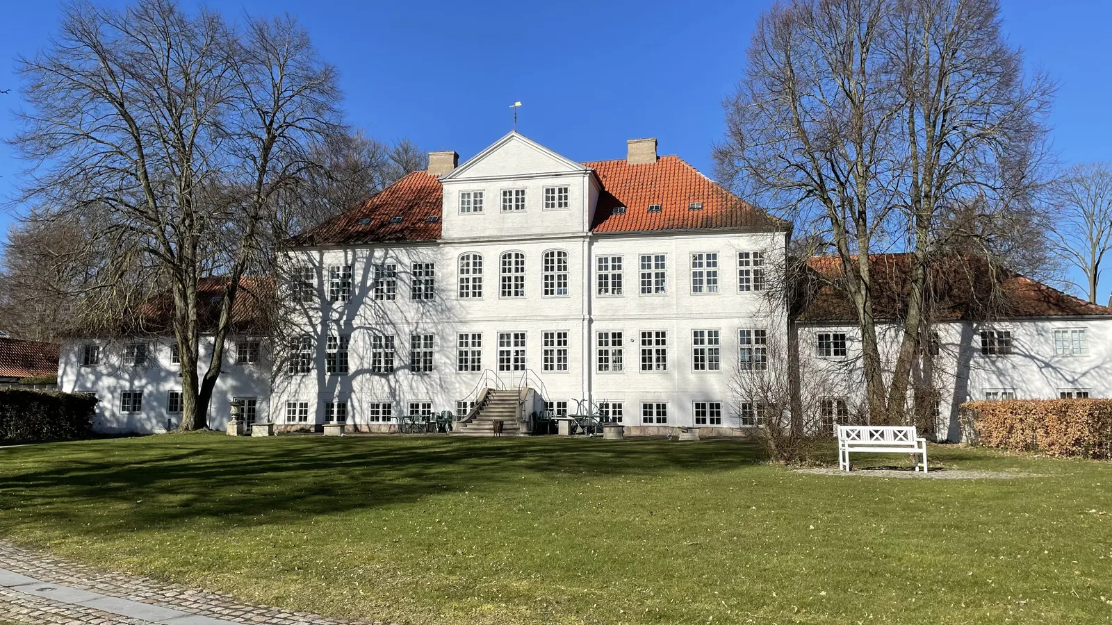 Historic white mansion with red roof, surrounded by trees on a sunny day. A cobblestone path leads through the green lawn in the foreground.