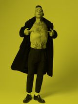 Man in a long coat and dress pants, revealing chest tattoos, standing against a yellow background.