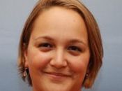 A headshot of Maha smiling gently against a slate blue background.