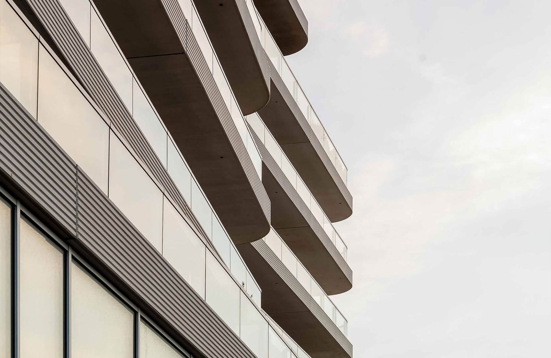 Modern building facade with glass and concrete balconies, featuring vertical and horizontal lines; sky in the background.