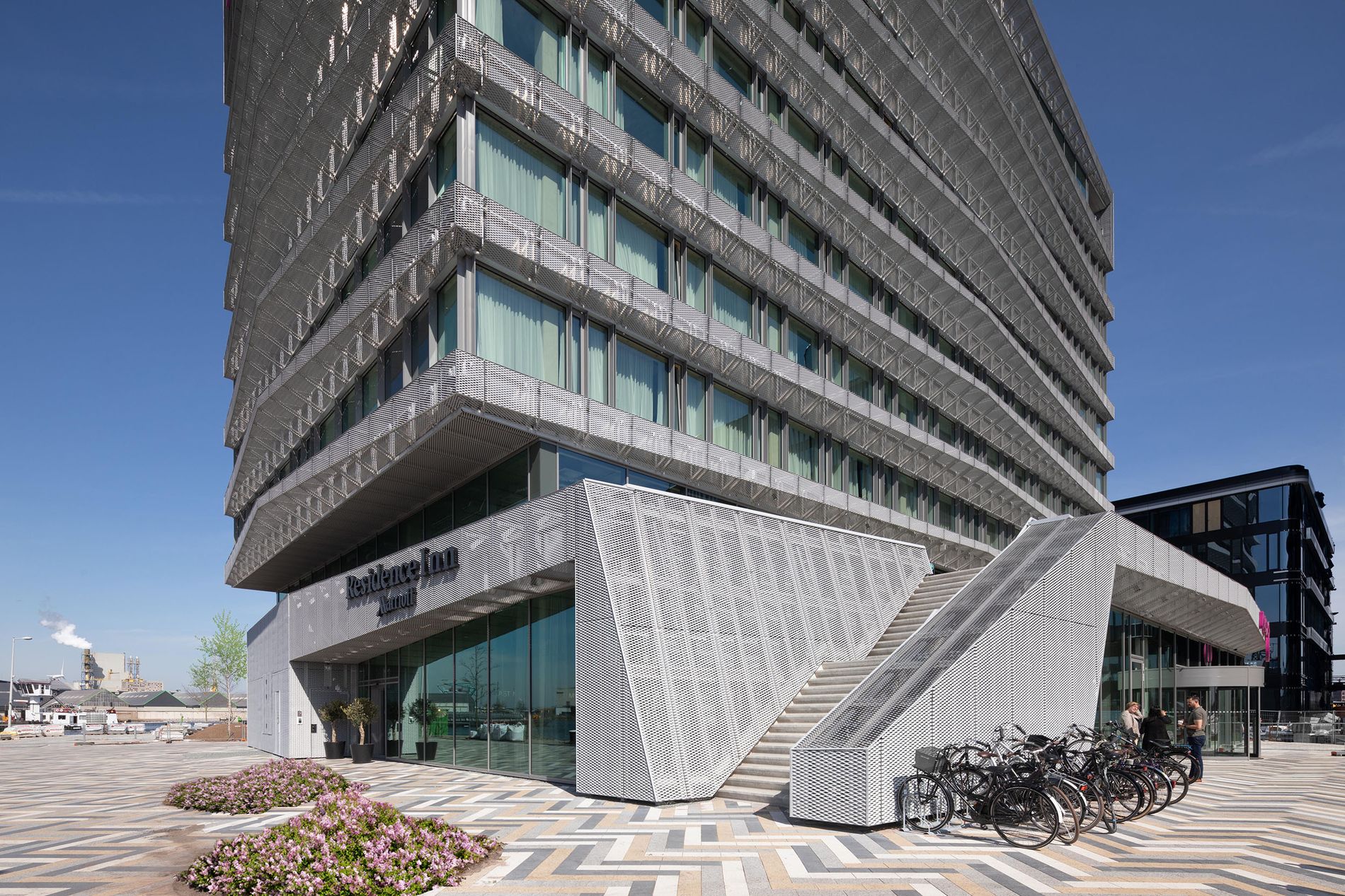 Modern hotel building with angular design, glass facade, outdoor stairs, and bicycles parked nearby under a clear blue sky.