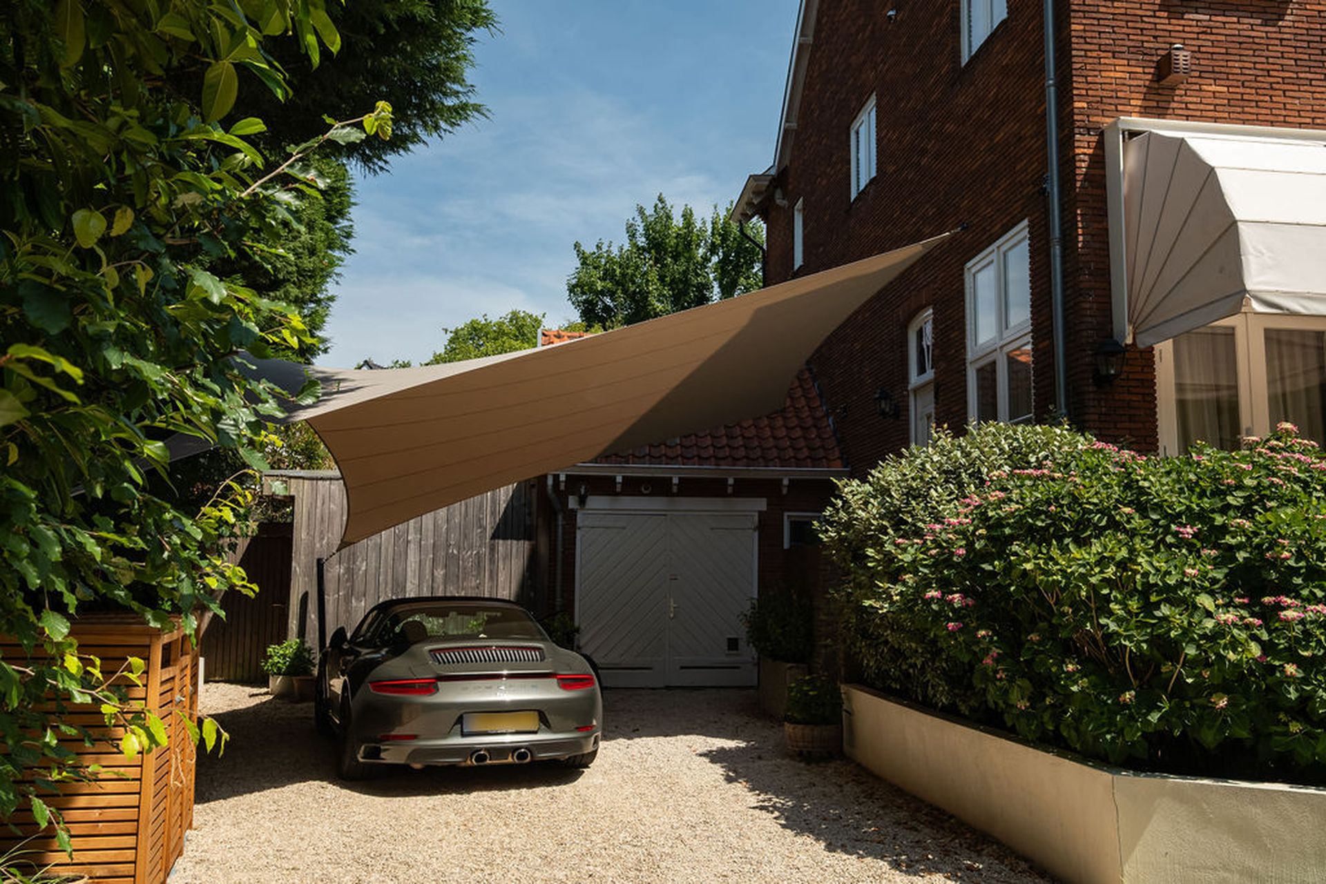 Sportauto geparkeerd op oprit naast huis met rode bakstenen, omgeven door begroeiing, onder een schaduwdoek.