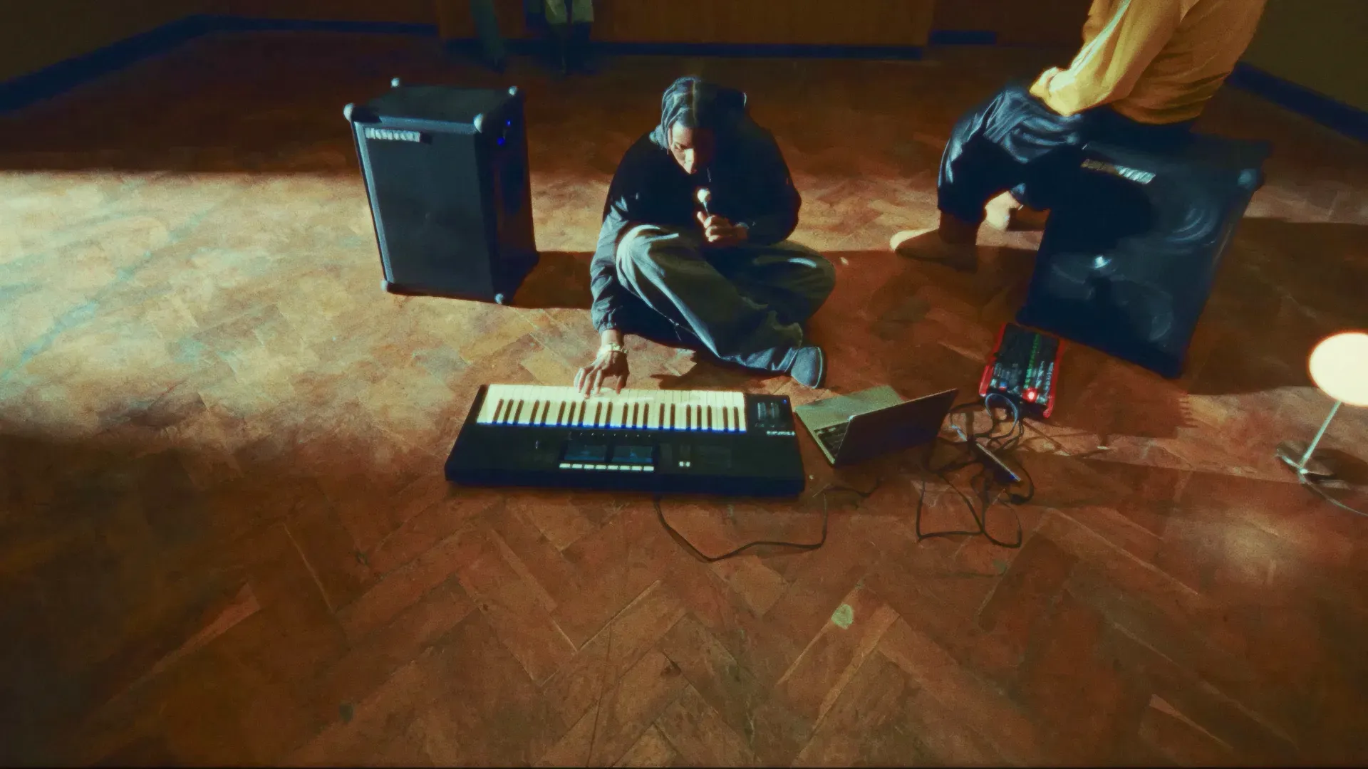Musician sitting on a parquet studio floor playing a keyboard between speakers, composing original music