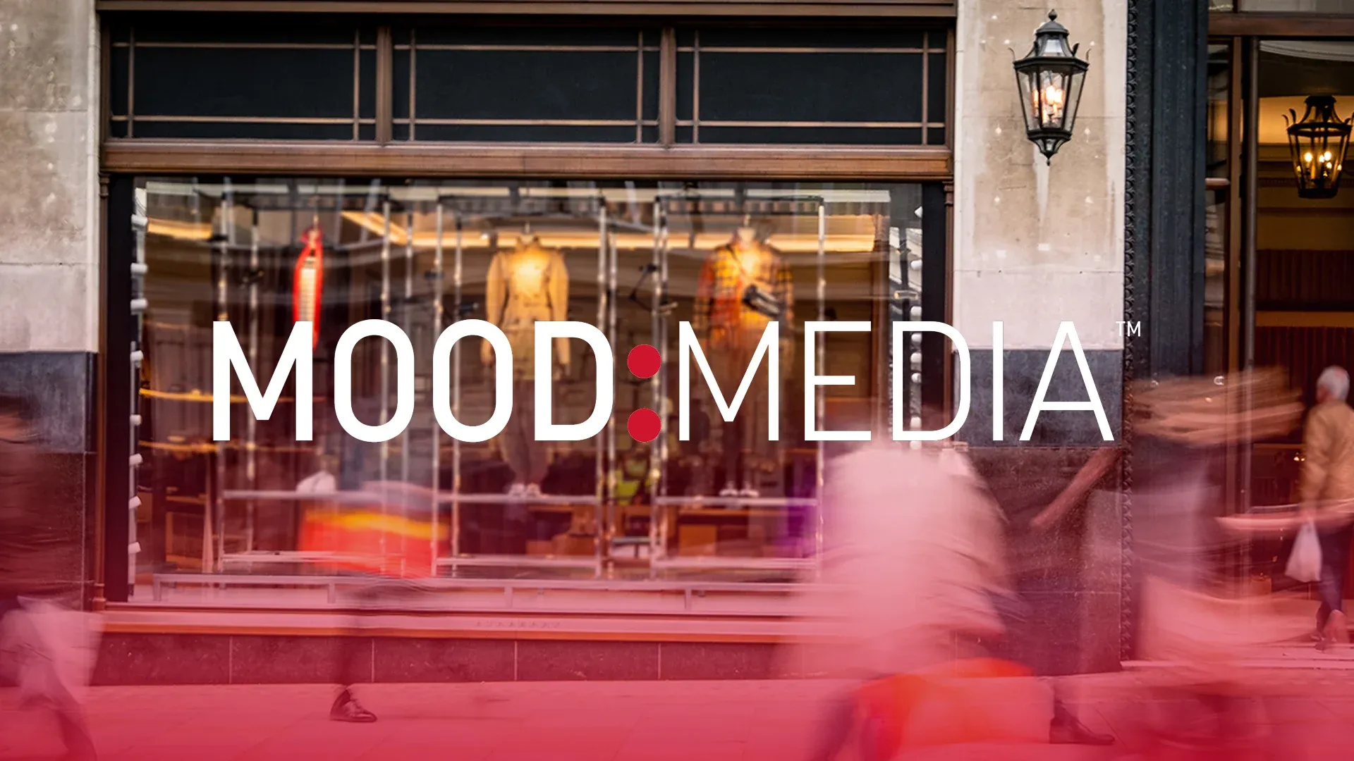 Wide photo of a retail storefront with the Mood Media wordmark while passersby walk past lit shop windows