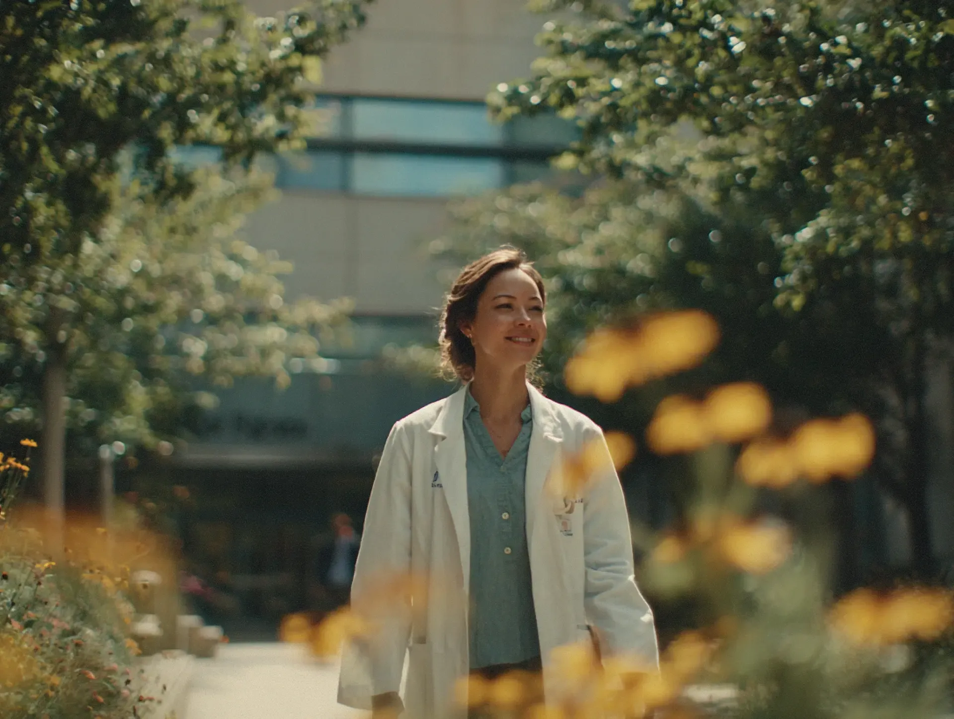 Clinician in a white coat walking through a sunlit hospital garden surrounded by trees and flowers