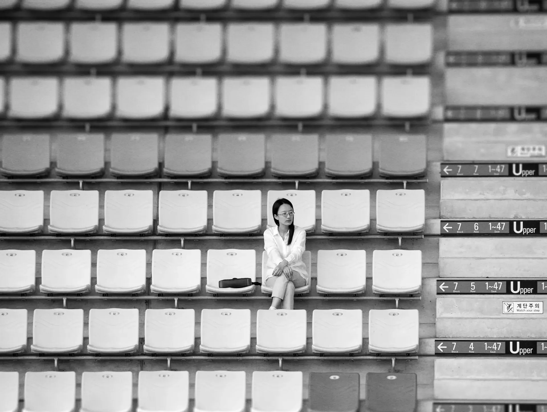 Black and white photo of a woman sitting alone among rows of empty stadium seats, gazing into the distance