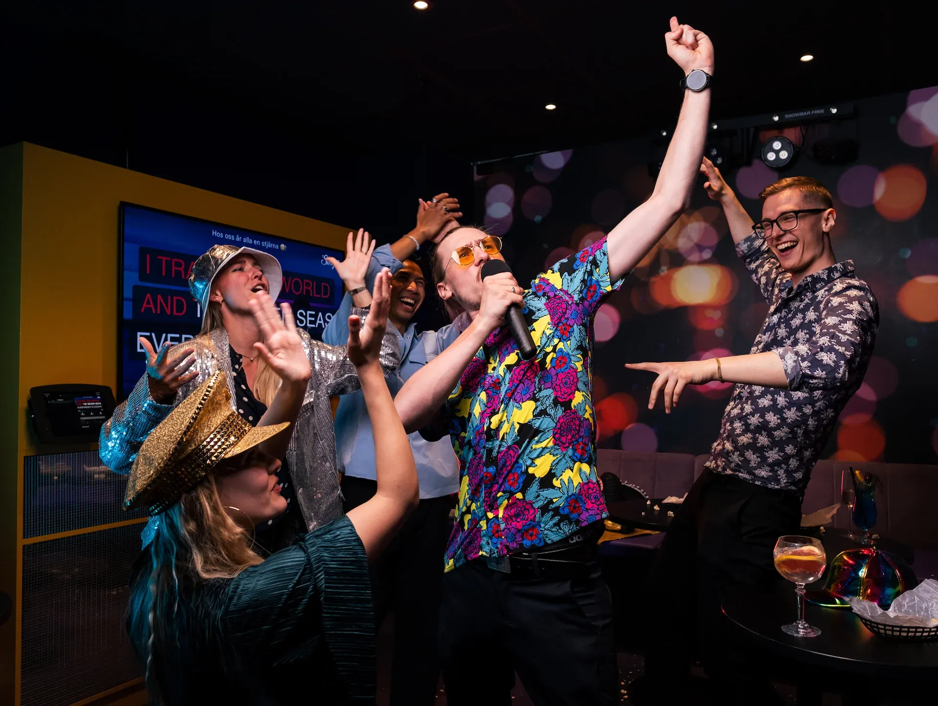 Group of friends celebrating and singing karaoke together in a private room with cocktails and a lyric screen