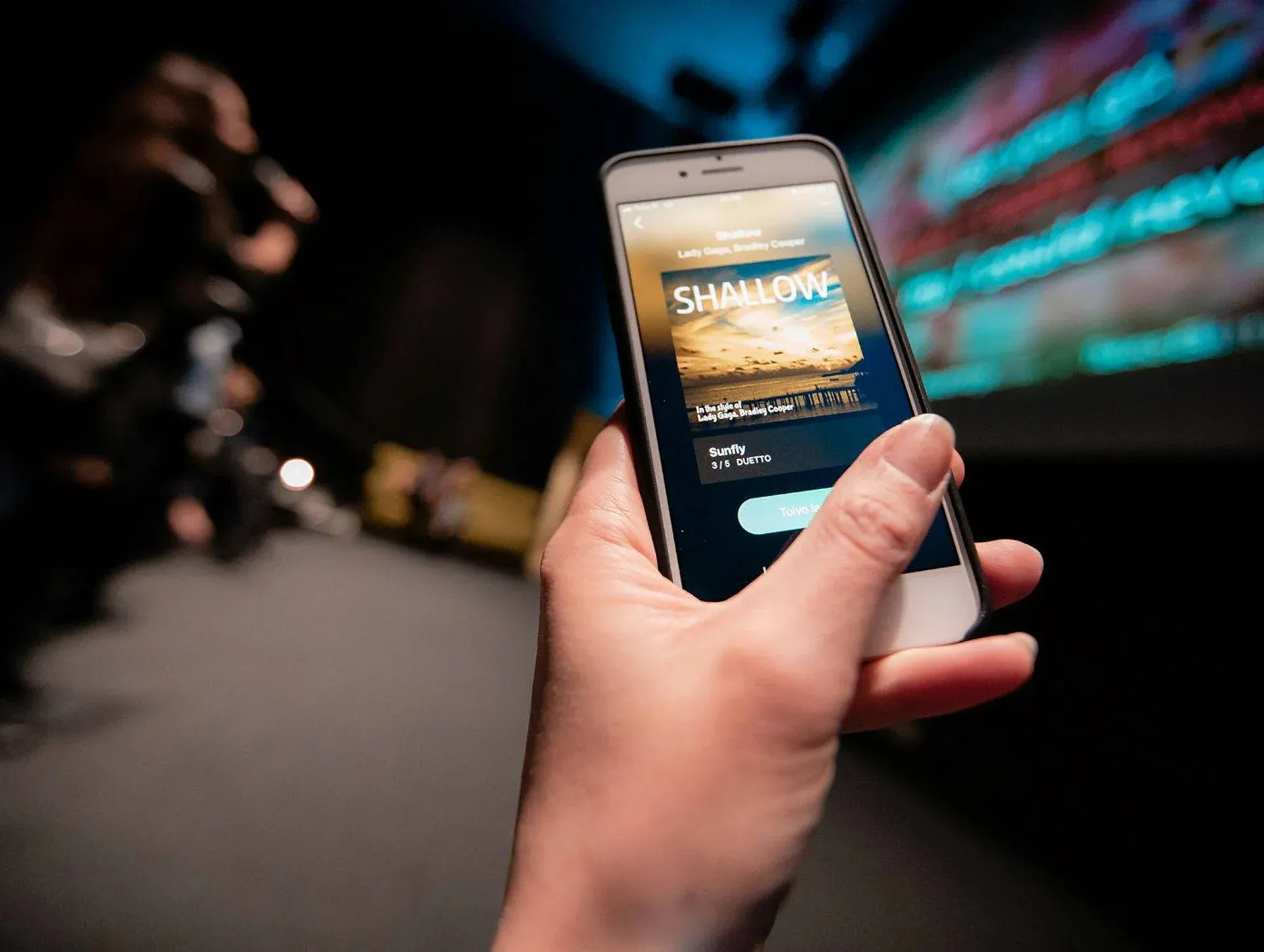 Hand holding a phone displaying a karaoke song selection screen with Shallow ready to play in a karaoke booth