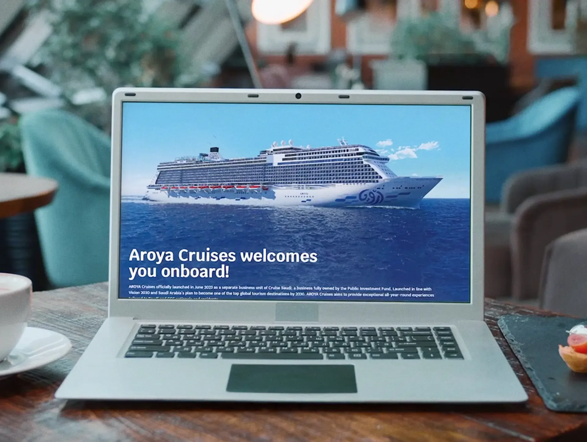 Laptop on a wooden table showing the Aroya Cruises welcome screen with a cruise ship, Sound of Aroya