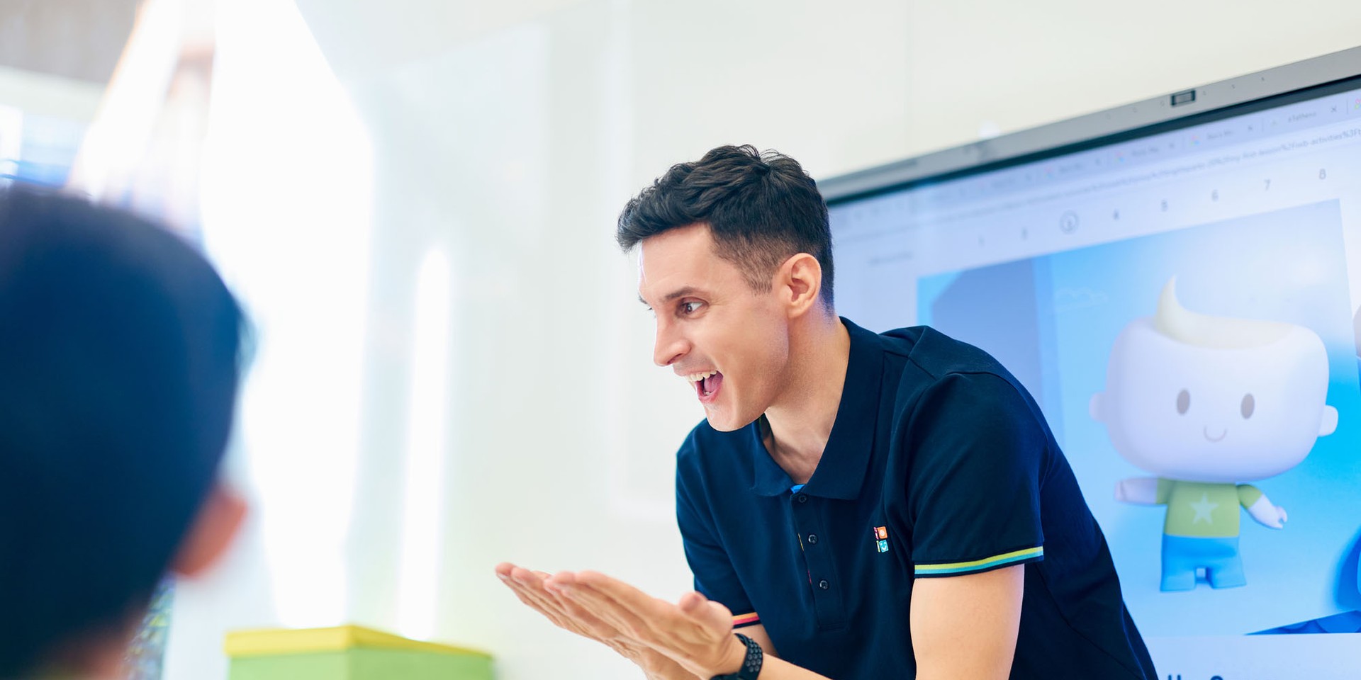Smiling man in a navy polo engages with a child, clapping hands in a classroom setting, with a cartoon character displayed on the screen behind