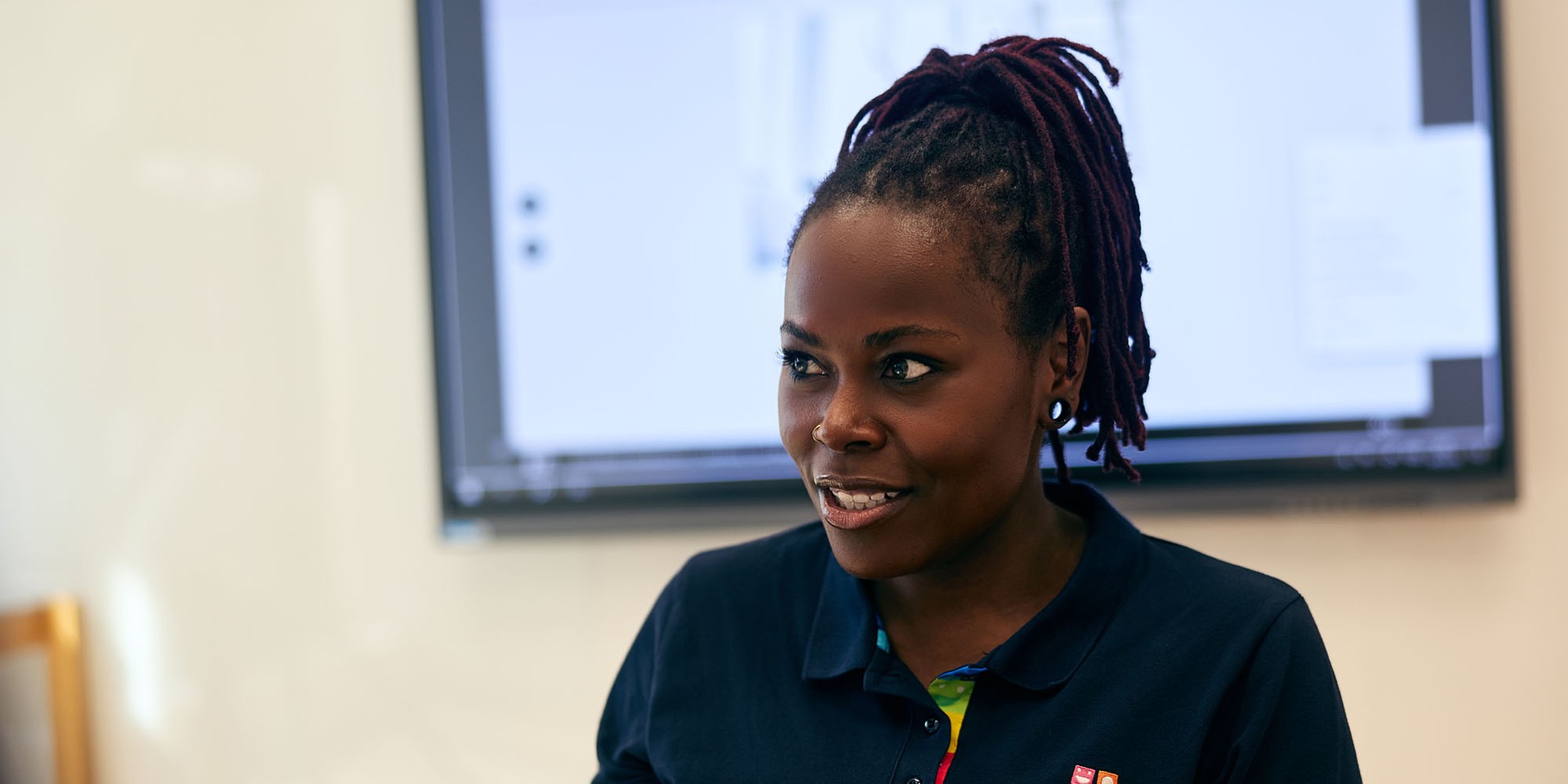 Person with dark hair in a ponytail, wearing a navy polo shirt with colorful details, stands in front of a screen, engaged in conversation.