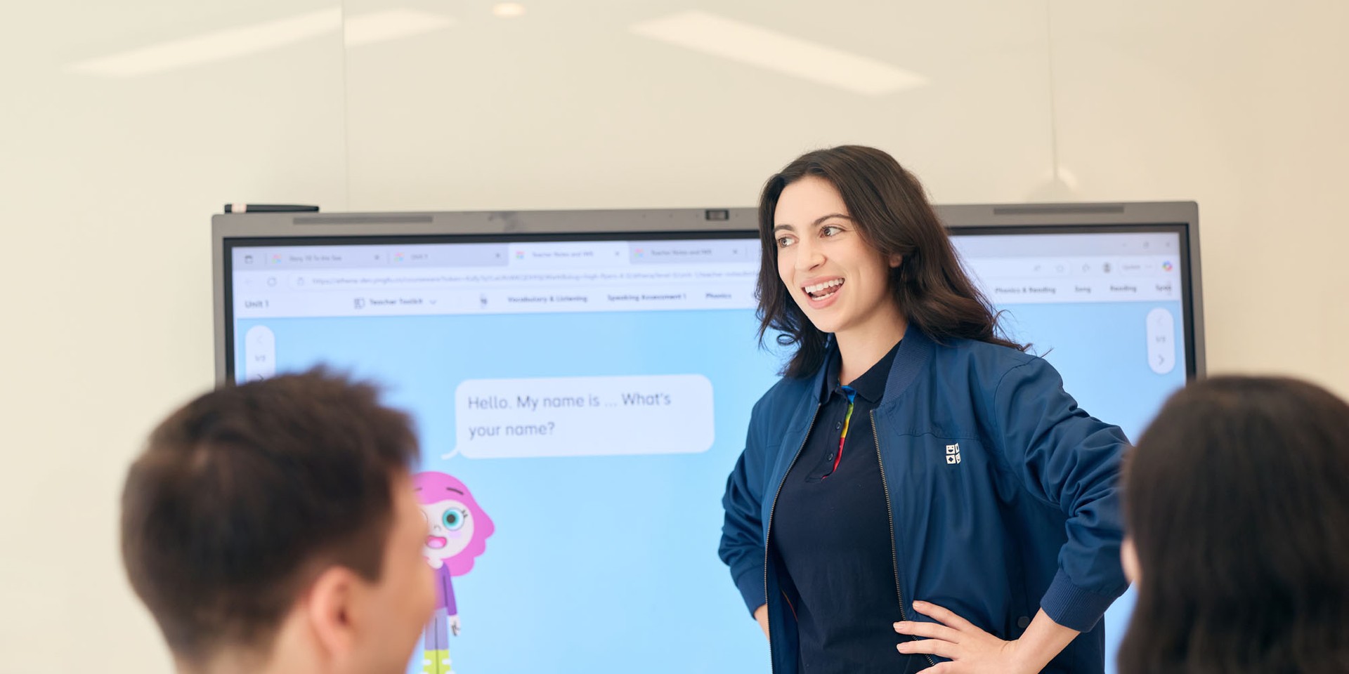 A woman stands smiling in front of a digital screen presenting to two seated individuals. The screen displays a playful character and text bubbles.