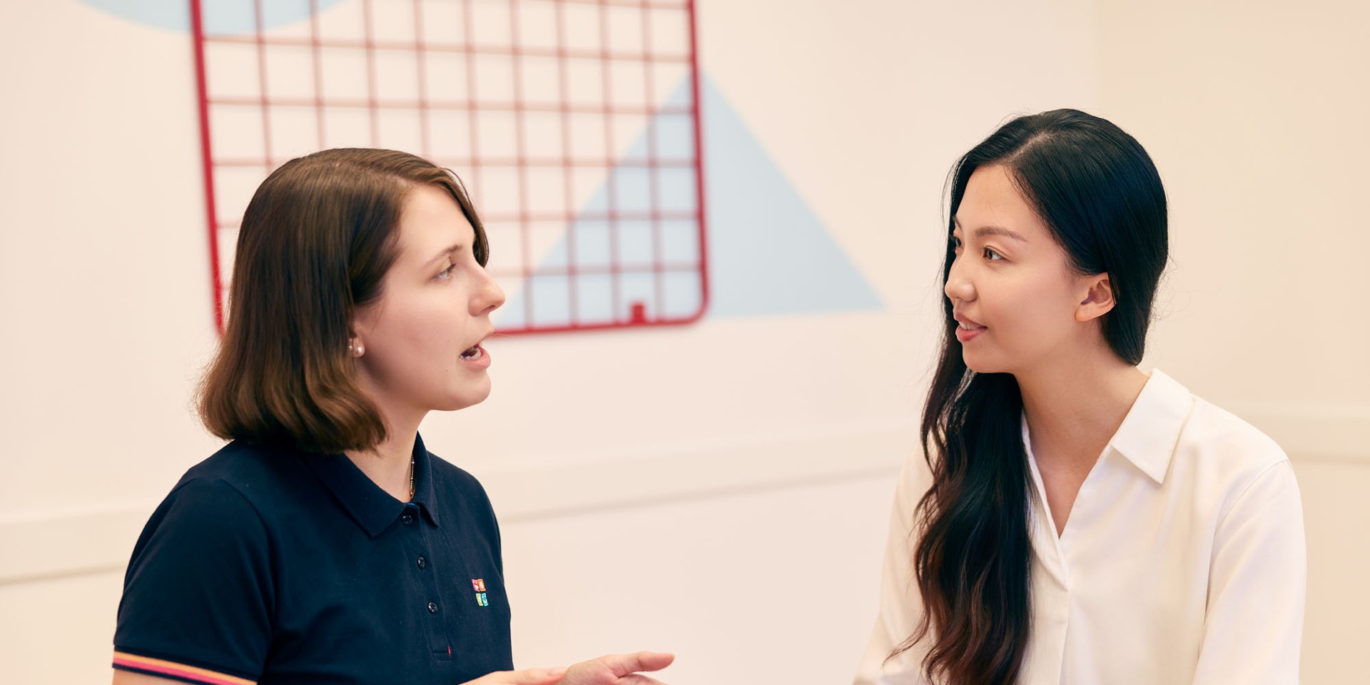 Two women sit facing each other and engage in a conversation in a classroom setting, one gesturing with her hands.