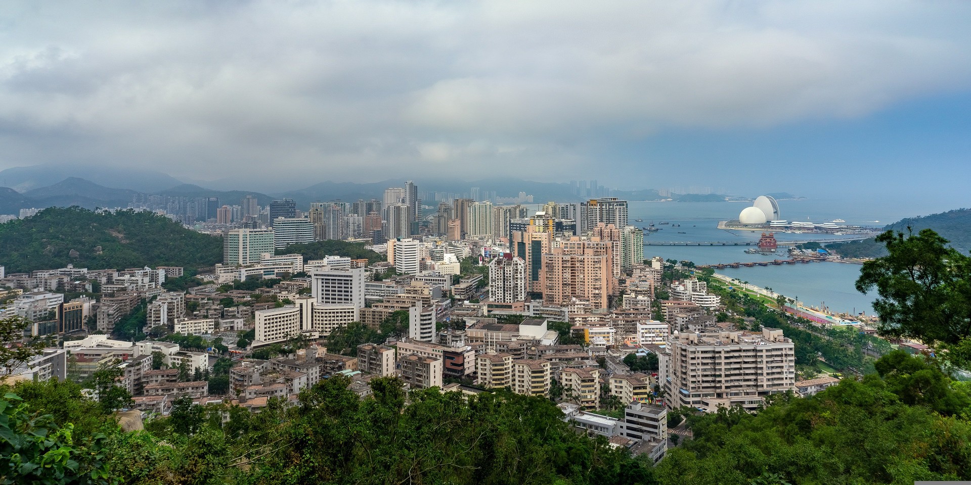 Aerial view of a coastal city with dense high-rise buildings, green hills, and a distinctive round white building by a bay under a cloudy sky.