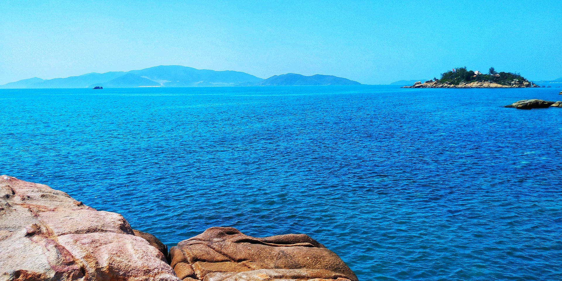 Alt Text Clear blue sea with distant islands on the horizon, rocks in the foreground, and a bright blue sky above.