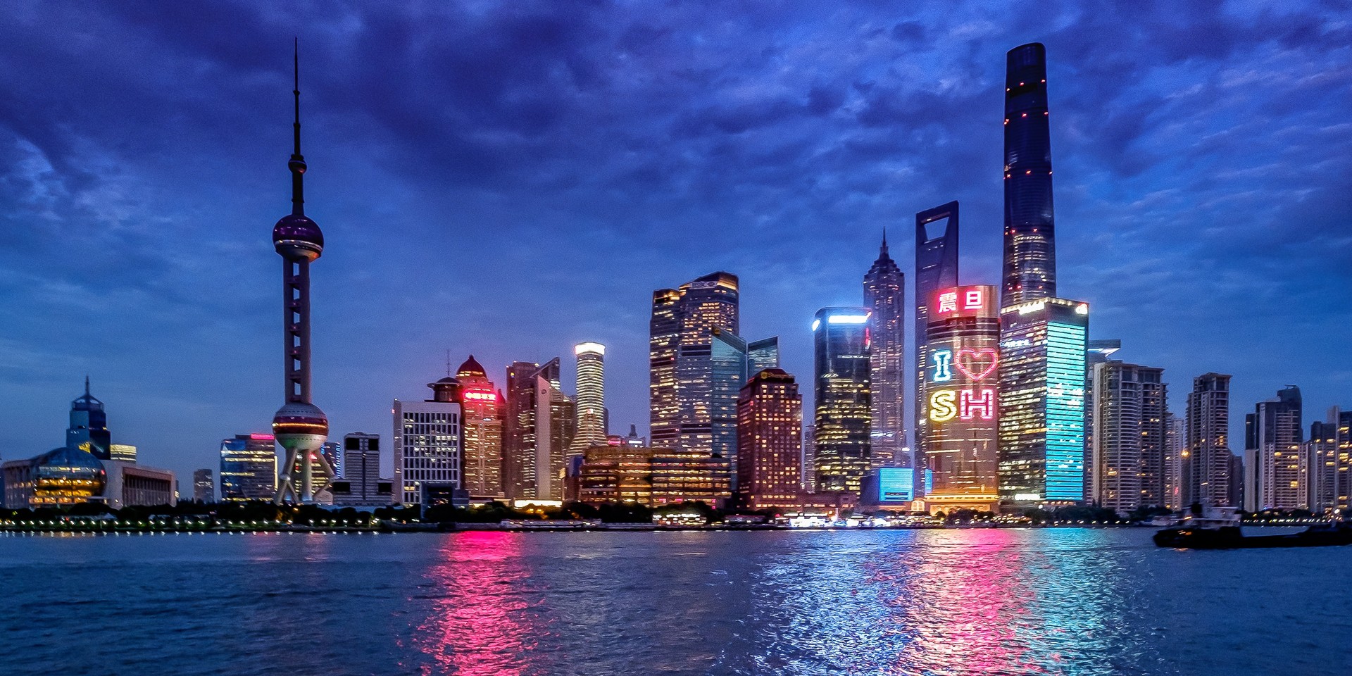 Shanghai skyline at night with illuminated skyscrapers, the Oriental Pearl Tower, and colorful reflections on the Huangpu River under a cloudy sky.