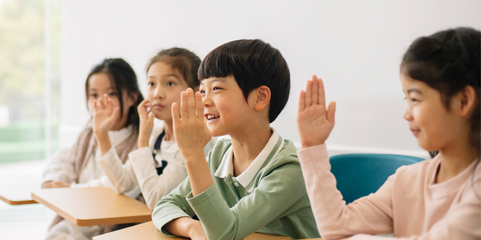 Four children seated at desks in a classroom, eagerly raising their hands, with focused expressions on their faces.